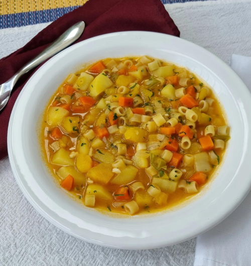 Tigela com sopa de legumes contendo massa ave-maria, cenoura, batata e vagem em caldo, servida sobre mesa clara.