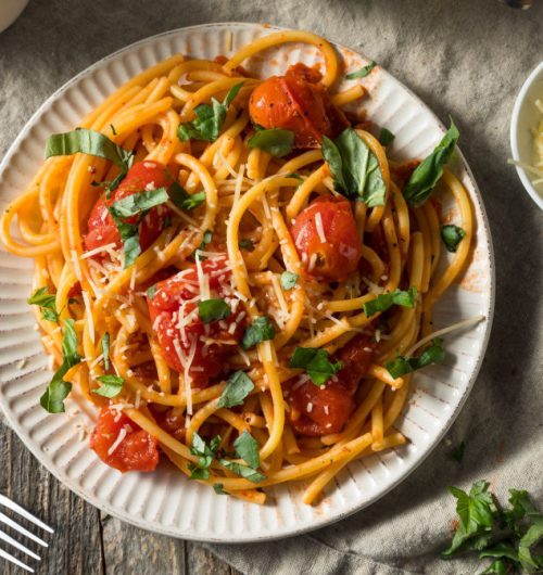 Prato de espaguete coberto com tomates, manjericão e queijo, disposto em prato branco sobre mesa clara.