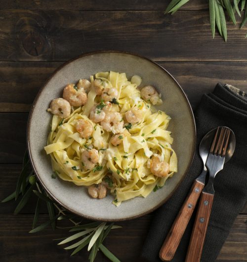 Prato de macarrão fettuccine servido com camarão, decorado com manjericão, em prato branco sobre mesa de madeira.