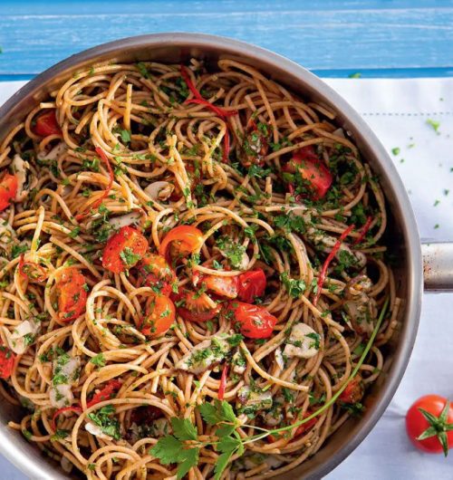 Prato de espaguete integral em panela funda, coberto com tomates, manjericão e champignon sobre mesa clara.