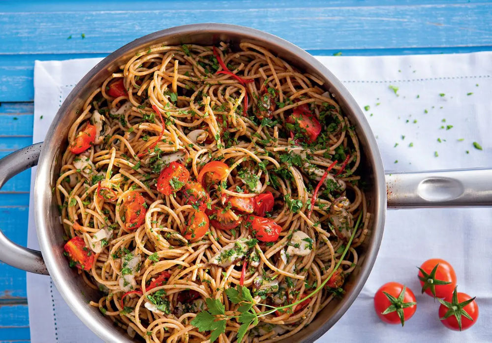 Prato de espaguete integral em panela funda, coberto com tomates, manjericão e champignon sobre mesa clara.