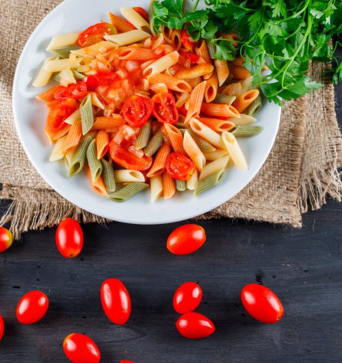 Prato de macarrão penne vegetias com molho de tomate e salsa, servido em prato branco, sobre mesa de madeira.