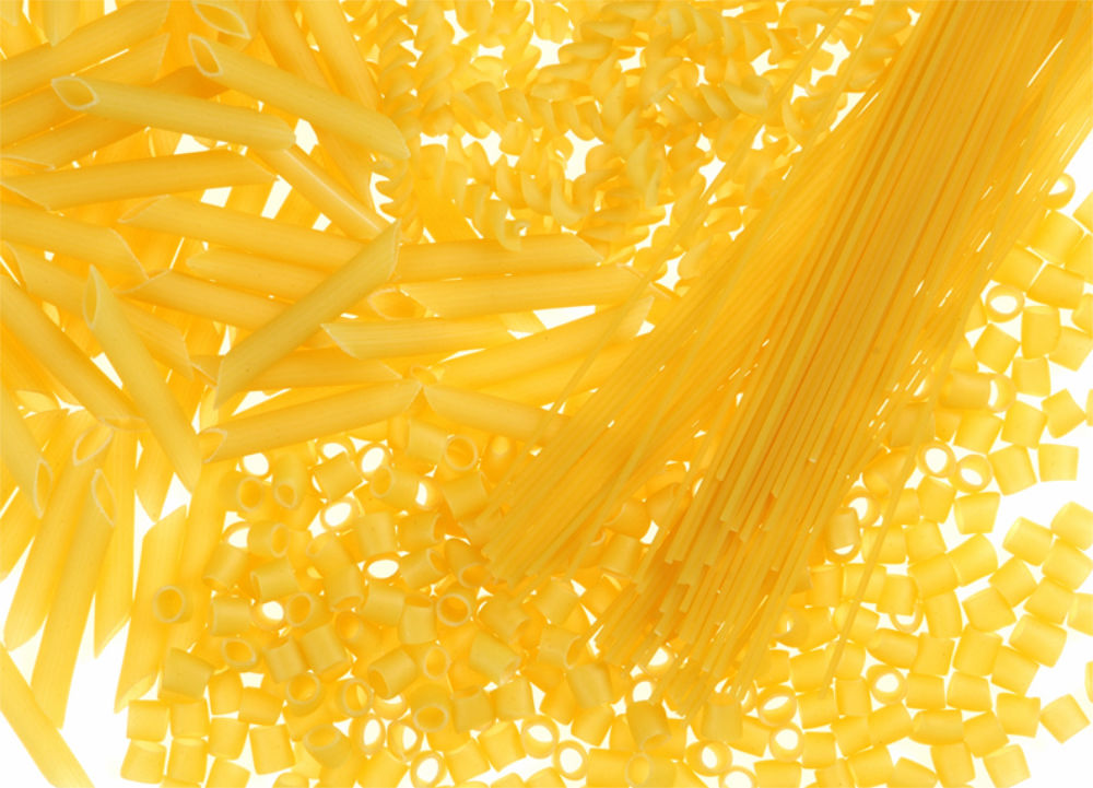 Variedade de massas cruas dispostas em superfície clara, incluindo espaguete, penne e fusilli.