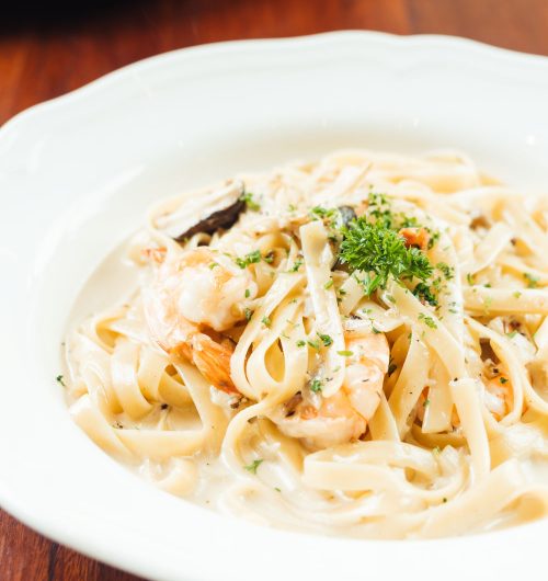 Prato de fettuccine ao molho branco com camarões, cogumelos e ervas, servido em prato branco, sobre mesa de madeira.