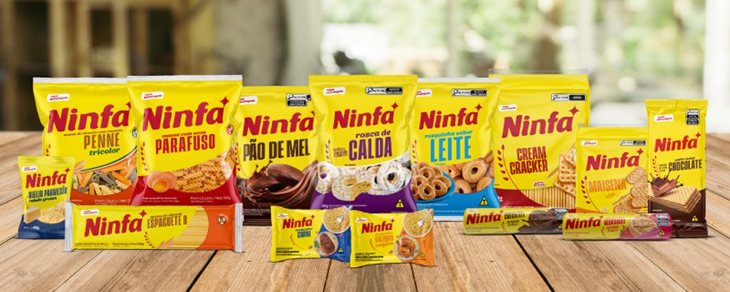 Diversas embalagens de produtos Ninfa, incluindo massas, biscoitos e wafers, dispostas lado a lado em pé, sobre fundo iluminado.