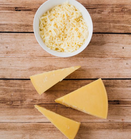 Queijo triangular em peça grande ao lado de porção de queijo ralado, dispostos sobre mesa de madeira.