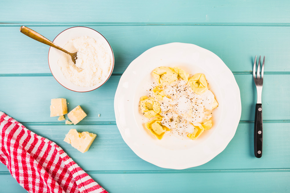 Prato de macarronada, polvilhado com queijo ralado fino, servido em prato branco sobre mesa azul.